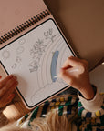 A child in a striped sweater colors a rainbow featuring winged animals, sun, clouds, and birds in the mushie Reusable Water Color Pad, using a refillable paintbrush for sustainable art fun.
