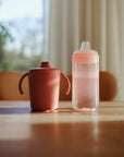 Two mushie Soft Spout Sippy Cups, including a pink one, sit on a sunlit table.