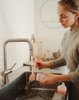 A woman in a gray sweater cleans a bottle at the sink with the mushie Silicone Baby Bottle Brush as water splashes. Another bottle with a pink lid sits on the counter nearby.