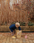 A young child in a blue sweater and yellow boots bends over a beige bucket outdoors, with tall dried grasses, a brick wall, and the mushie Water Wheel Bath Toy next to a pink watering can in the background.