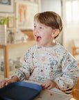 A young child with light brown hair, wearing a colorful bib, smiles while sitting at a table with mushie's Square Dinnerware Plates on a blue tray in a bright, cozy room.