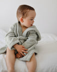 A toddler in a mushie Bear Robe sits on a bed, looking to the side against a white background.