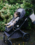 A toddler with a mushie Adjustable Silicone Strap 2-Pack sits in a gray stroller near green bushes.