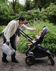 Woman hands baby a mushie Adjustable Silicone Strap, both smiling together outdoors in the park.