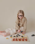 A young child with long blonde hair and glasses sits on the floor, smiling while playing with the Mushie Wooden Puzzle—an educational toy designed to encourage fine motor skills—on a beige background.
