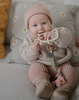 A baby in a pink knit hat and outfit sits on a couch, holding and chewing on a FRIGG Butterfly Anatomical Silicone Pacifier (6-18 Months). Pillows, including one with daisies, and a pink blanket are in the background.