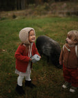 Two young children in warm autumn clothes smile at each other on the grass. One holds a toy and the mushie Silicone Pacifier Clip | Hera, while a black pig grazes nearby among trees and a fence in the blurry minimalist background.