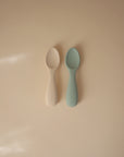 Two small, rounded mushie Silicone Toddler Starter Spoons—one off-white, one pale teal—lie side by side on a beige surface. Embossed with the mushie logo, they're perfect for encouraging independent feeding in toddlers.
