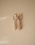 The mushie Silicone Toddler Starter Spoons come in light beige and light brown, with "mushie" embossed on each handle. Two spoons are shown side by side on a smooth, neutral surface—perfect as starter spoons for toddlers.