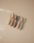 The mushie Silicone Toddler Starter Spoons set includes five pastel-colored spoons (cream, beige, blue, pink, green) arranged neatly on a light beige surface—ideal for encouraging independent feeding for toddlers.