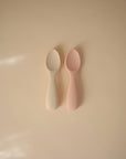Two mushie Silicone Toddler Starter Spoons, one off-white and one light pink, rest side by side on a beige surface. Both have the brand name embossed on their handles, ideal for encouraging independent feeding. Soft light casts gentle shadows nearby.