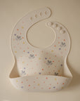 The mushie Bluey x Mushie Silicone Baby Bib, featuring small stars, hearts, and Bluey-inspired animal illustrations with a pocket, is laid flat on a light beige background.