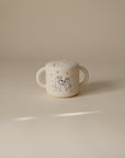 The mushie Bluey x Mushie Snack Cup is a baby feeding essential featuring two-handle design and a playful cartoon of two smiling cats in hats with stars on a plain light background.
