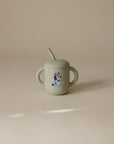 The Mushie Bluey x Mushie Silicone Training Cup + Straw is a light gray, food-grade silicone cup with two handles and a straw, featuring a cartoon Bluey dog and white stars on the front, shown on a plain beige background.