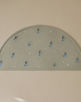 The mushie Bluey x Mushie Silicone Placemat is light grey, semicircular, and features small Bluey characters with scattered white stars. The brand name is embossed in one corner and it rests on a beige surface.