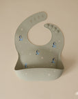 The Bluey x Mushie Silicone Baby Bib by mushie features a light green color, a pocket, and playful cartoon dogs with white stars—ideal for fans of Mushie bibs and cute silicone baby accessories.