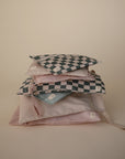 A stack of pastel mushie Water Resistant Wet Bags, including two with blue and white checkerboard patterns, displayed against a beige background. Each bag features a zipper, diaper bag storage design, and a small white label.