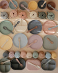 A neatly arranged grid of mushie Square Dinnerware Plates, BPA-free silicone in pastel colors, alongside matching bowls, cups, forks, spoons, and snack containers in various shapes on a light surface.
