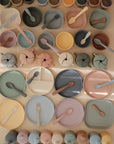 Pastel-colored mushie Silicone Toddler Starter Spoons are neatly arranged by color and type on a light surface, creating an organized display. Ideal for independent feeding, these silicone spoons are perfect for toddlers.