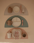 Three mushie Silicone Placemat sets in beige, green, and pink are shown, each with a matching cup, bowl or plate, utensils, and accessories—all made from food-grade silicone for children.