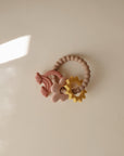 The mushie Nature Teething Ring is a beaded sensory baby toy and silicone teether featuring cream, pink, and yellow shapes—a rainbow, flower, and sun—arranged on a beige surface with soft light and gentle shadows.