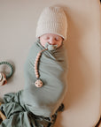 A newborn baby swaddled in a sage green blanket lies on a beige surface, wearing a knit hat and using a pacifier attached to a beaded clip. A teething ring is placed nearby.