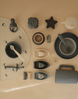 A flat lay of Mushie Classic Silicone Suction Plate and other neutral-toned silicone baby feeding items, spoons, cups, animal print placemat, stacking toys, star teether, and boat toys arranged on a beige background.