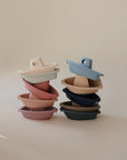 Two stacks of mushie Bath Boats, made from non-toxic plastic in pastel colors, are arranged against a neutral background. The left stack features pink and beige boats, while the right showcases blue and navy water play toys.