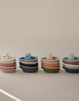Four stacks of mushie Bath Boats in non-toxic plastic, each with handles and spouts. Shown in muted pastel and earthy colors on a neutral background. Perfect for water play or as stackable bath toys.