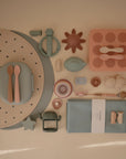 Flat lay of mushie baby feeding and teething essentials—featuring food-grade silicone spoons, divided plate, bib, teething rings, molds, cup, bowl, napkins, and a Silicone Placemat—all neatly arranged on a beige surface.