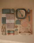 A flat lay of mushie’s pastel-colored BPA-free Square Dinnerware Plates, alongside a bib, bowl with spoon, silicone pacifier, cloths, teether, and a rainbow-shaped accessory arranged neatly on a light beige surface.
