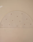 The mushie Silicone Placemat is a semicircular, light-colored food-grade silicone mat featuring scattered pretzel illustrations and an embossed mushie logo in the lower right corner, set against a plain background—ideal for fans of the brand.