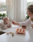 A woman smiles as she feeds a baby in a high chair using mushie Silicone Toddler Starter Spoons. A small bowl and pacifier are on the table, sunlight streams through white-curtained windows, and green plants sit on the sill.