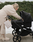 A woman in a beige coat and boots leans over a black stroller decorated with mushie Chain Link Rings on a snowy sidewalk, with a hedge in the background.