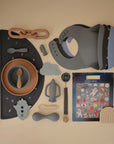 A flat lay of mushie baby feeding items in soft colors, featuring a bib, bowl, Silicone Toddler Starter Spoons, fork, teether, rocket toy, placemat, and a space-themed sticker sheet on beige—ideal for independent feeding.