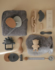 A flat lay of baby essentials in neutral tones, featuring mushie Cradle Cap Brushes, a gray towel, wooden brush, teether, toy car, lotion, spoons, pacifier clip, teething ring, and silicone toys neatly arranged on a beige background.