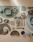 A flat lay of mushie silicone Toddler Starter Spoons, plates, bowls, bibs, teethers, stacking toys, and a rainbow in soft green, taupe, and gray arranged on a beige background.