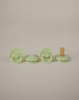 Two light green FRIGG Rope Silicone Pacifiers by FRIGG, crafted from medical-grade silicone and featuring unique rope designs, are lined up on a neutral background—perfect essentials for your baby's comfort.
