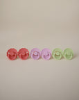 Four FRIGG Daisy Natural Rubber Pacifiers in pink, purple, and light green are lined up on a light background; each features a natural rubber latex nipple for comfort. Available in the FRIGG Daisy Natural Rubber Pacifier 2-Pack.