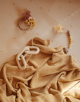 A soft beige blanket with textured dots is spread out, topped with a cream-colored teething toy. Nearby, a pacifier and a pastel beaded ring toy rest on a crib sheet decorated with small yellow stars.