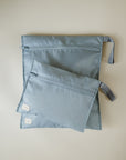 Two light blue mushie Water Resistant Wet Bags with small white tags and side straps rest on a light surface. The smaller pouch partially overlaps the larger, ideal for diaper bag storage.
