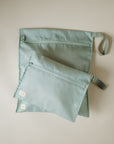 Two light green, rectangular mushie Water Resistant Wet Bags with zippers rest on a light surface. The larger bag has a side loop, while the smaller slightly overlaps it—ideal for compact diaper bag storage.