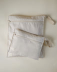 Two light beige mushie Water Resistant Wet Bags with handles are displayed flat on a light surface. The larger bag is beneath a smaller pouch, both featuring small white tags in the lower left corner for easy storage.