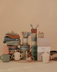 A set of Mushie pastel baby feeding essentials—cups, bowls, utensils, bibs, pacifiers, stackable containers, a packaged muslin cloth, and the Splat Mat—are neatly arranged against a beige background.