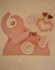 Pink mushie Silicone Baby Bib set with a placemat, bowl, spoon, cup, and toys.