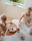 Two young children sit at a marble table set for a meal, each with a Mushie Classic Silicone Suction Plate. The table features woven holders and a flower-shaped accent as sunlight streams in through the window behind them.