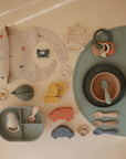 The Mushie Classic Silicone Suction Plate set, displayed on a beige surface, includes non-toxic plates, bowls, utensils, teether, pacifier, toy vehicles, and bib in soft pastels—perfect for safe and playful toddler feeding.