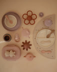A flat lay of mushie pastel-colored silicone Lunchboxes and matching feeding items, including plates with compartments, bowls, cups, spoons, a bib, teether, and placemat, all neatly arranged on a beige background.