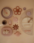 A flat lay of mushie Silicone Toddler Starter Spoons and matching pastel feeding items—plates, bowls, cups, placemat, teether, and snack container—perfectly arranged to support independent feeding on a beige background.