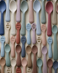 A flat lay of Mushie First Feeding Baby Spoons in navy, sage, blush, mauve, rust, and blue-grey, arranged on a neutral background—ideal for supporting your baby's self-feeding journey.